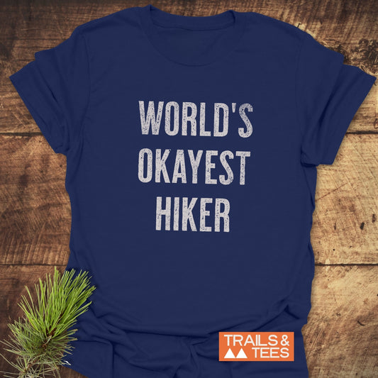 Navy-blue T-shirt with WORLD’S OKAYEST HIKER in white font, featuring TRAILS & TEES logo, laid on a wooden surface with a pine sprig for decoration.