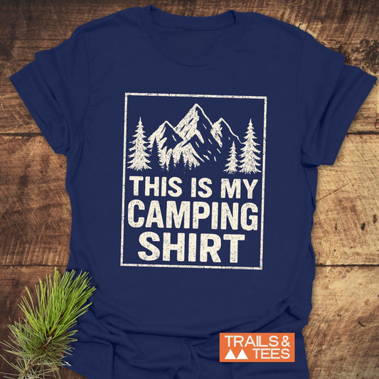 Navy-blue This Is My CAMPING T-Shirt with white print of pine trees and mountains, laid on wood with a pine sprig. Features include soft cotton and durable print.