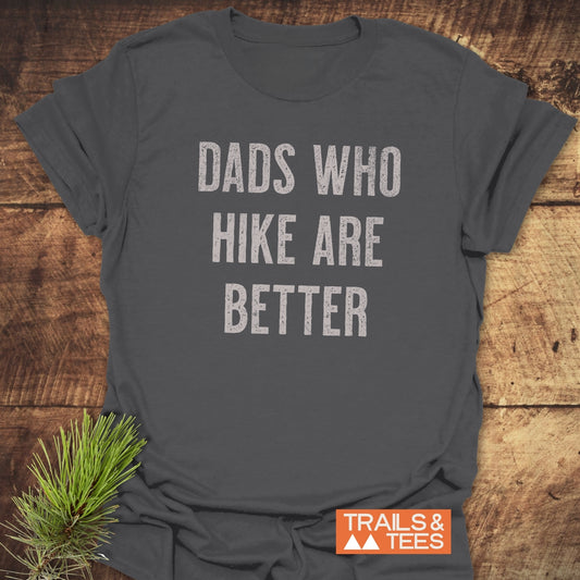 Dark-gray Dads Who Hike Are Better T-shirt on wood, featuring distressed light lettering, small orange Trails & Tees logo, and pine sprig accent.