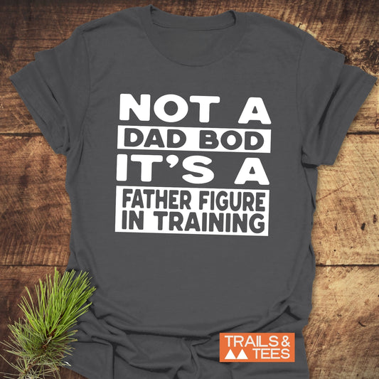 It's Not A Dad Bod T-Shirt with bold white text, laid on wood. Soft, ring-spun cotton, durable print, orange TRAILS & TEES logo visible.