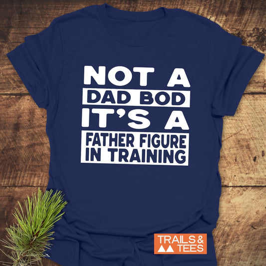 It's Not A Dad Bod T-Shirt with bold white text on navy-blue, featuring durable prints. Laid on wood with pine sprig, Trails & Tees logo visible.