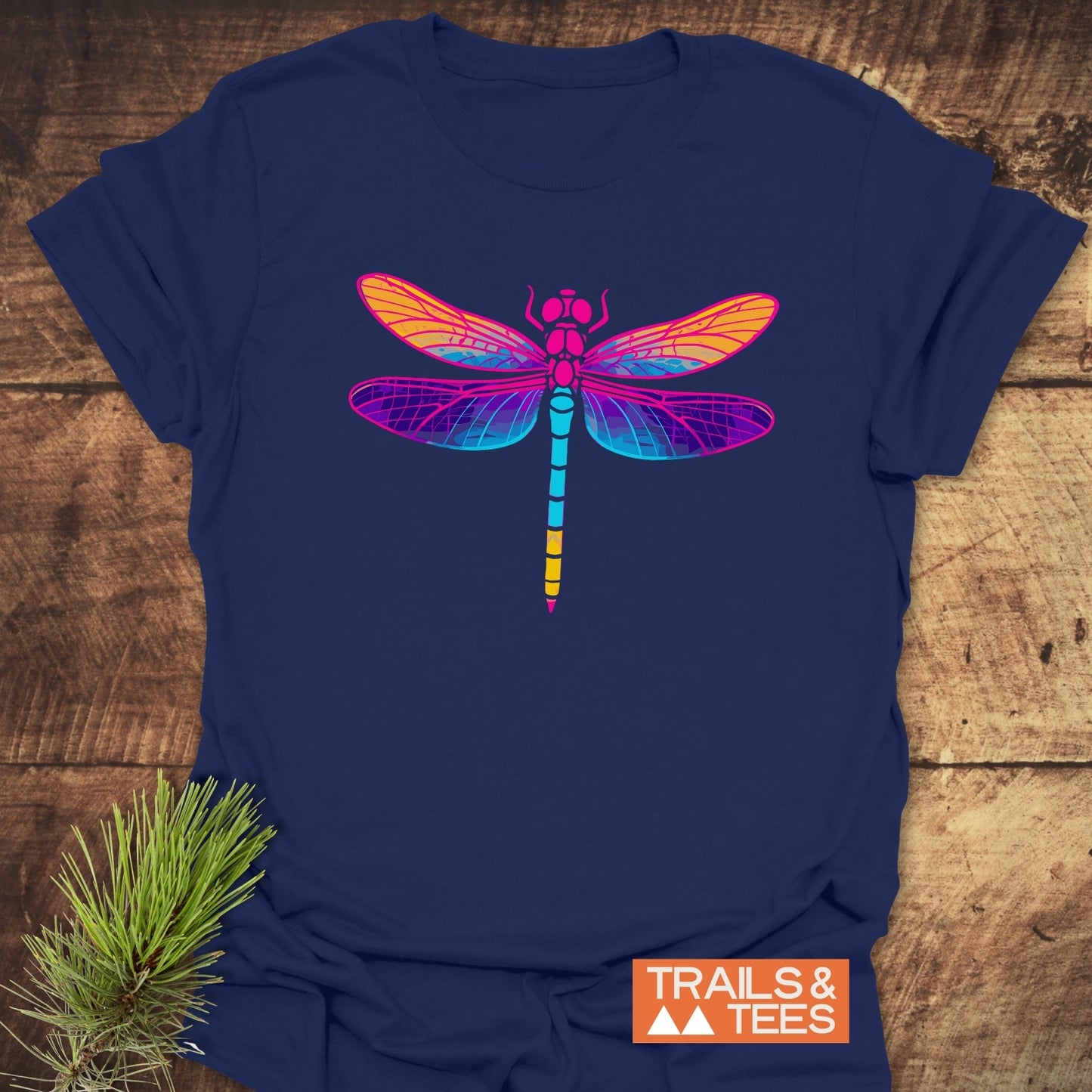 Psychedelic Dragonfly T-Shirt on wooden surface, featuring a vibrant dragonfly design. Made from soft, durable cotton for outdoor adventures. TRAILS & TEES logo visible.