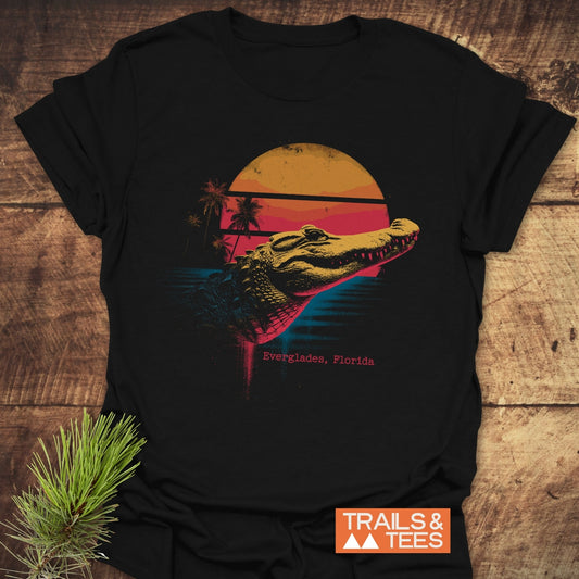 Everglades Alligator T-Shirt featuring a retro alligator graphic with palm trees and sun, laid on wood. Highlights comfort, durability, and eco-friendly print.