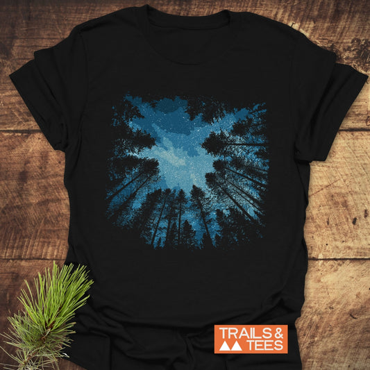 Starry Night Forest T-Shirt featuring a night-sky scene with tree silhouettes, crafted from soft cotton, laid on wood with a pine sprig and TRAILS & TEES logo.