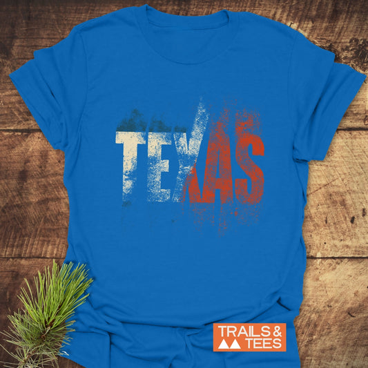 TEXAS T-Shirt on wooden surface, featuring a distressed print and TRAILS & TEES logo, styled with a pine sprig, showcasing comfort and outdoor spirit.