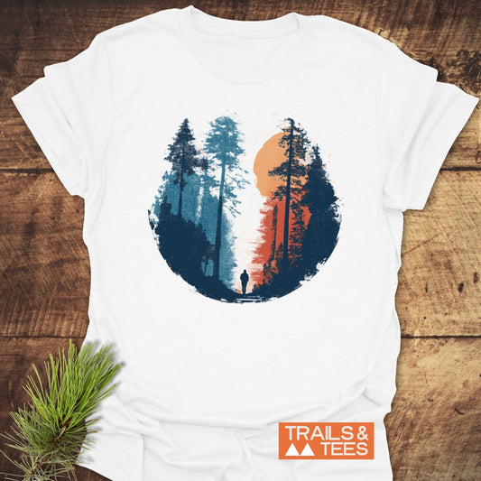 Sunset Hiker T-Shirt with a forest scene graphic in blue and orange tones, featuring a silhouette and branded TRAILS & TEES label, laid on wood.