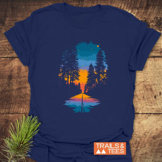 Into The Light T-Shirt: Navy-blue tee with a forest sunset graphic, featuring pine trees and a water reflection. Includes TRAILS & TEES logo, evergreen sprig accent.