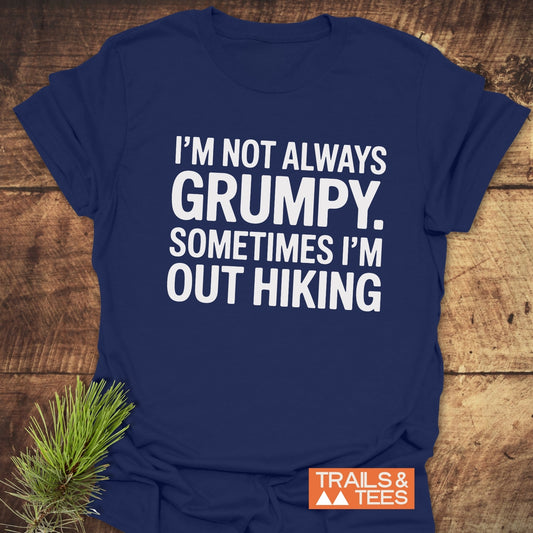 Im Not Always Grumpy T-Shirt on wooden surface, featuring bold white text and Trails & Tees logo. Pine sprig beside it enhances the outdoor theme.