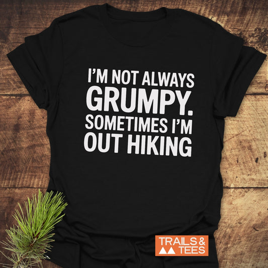 Black T-shirt with “I’M NOT ALWAYS GRUMPY. SOMETIMES I’M OUT HIKING” in white text, styled with pine sprig, featuring Trails & Tees logo.