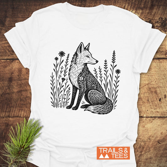 Wildflower Fox T-Shirt featuring a hand-drawn fox amid grasses and wildflowers, laid flat on wood with TRAILS & TEES logo visible.