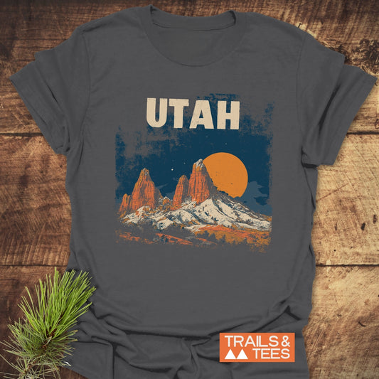 Utah Retro T-Shirt with mountain scene graphic, folded on wood surface with pine sprig. Made from soft cotton, ideal for outdoor adventures.