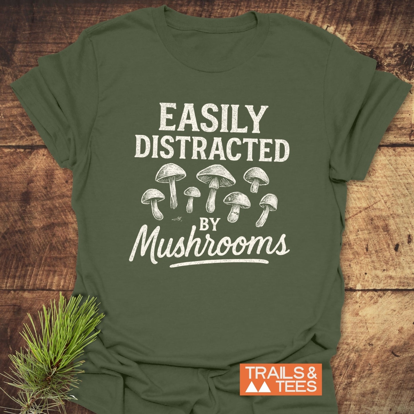 Easily Distracted By Mushrooms T-Shirt with line-drawn mushrooms, in forest-green cotton, laid on wood surface. Features durable, pre-shrunk fit and eco-friendly printing.