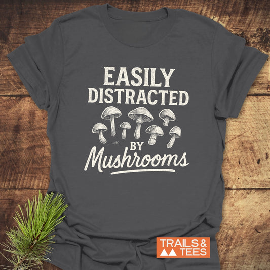 Easily Distracted By Mushrooms T-Shirt with a hand-drawn mushroom design, on a wooden surface. Includes “TRAILS & TEES” logo, emphasizing outdoor adventure and comfort.