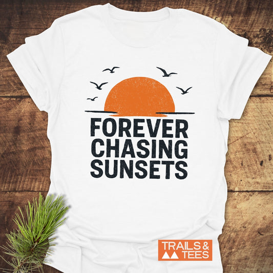Forever Chasing Sunsets T-Shirt with an orange half-sun and birds graphic, featuring bold slogan and Trails & Tees logo, displayed on a wooden surface.