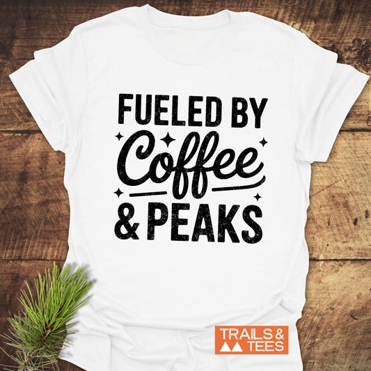 Fueled By Coffee And Peaks T-Shirt on rustic wood, features black lettering, pine sprig accent, and Trails & Tees logo, embodying outdoor adventure spirit.