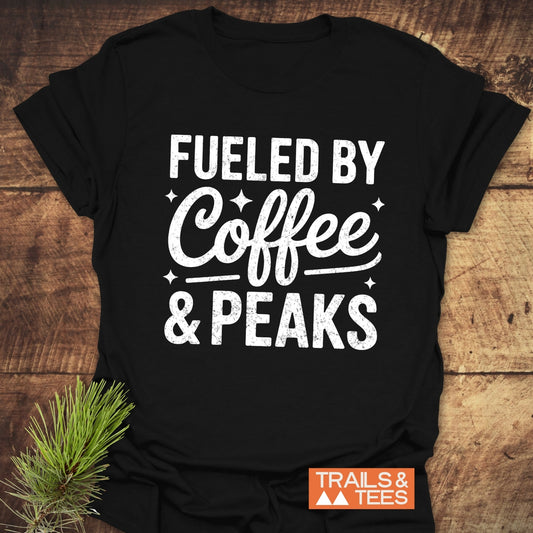 Fueled By Coffee And Peaks T-Shirt on wood surface, features bold slogan, FUELED BY Coffee & PEAKS, in white, distressed font. Trails & Tees brand mark visible.