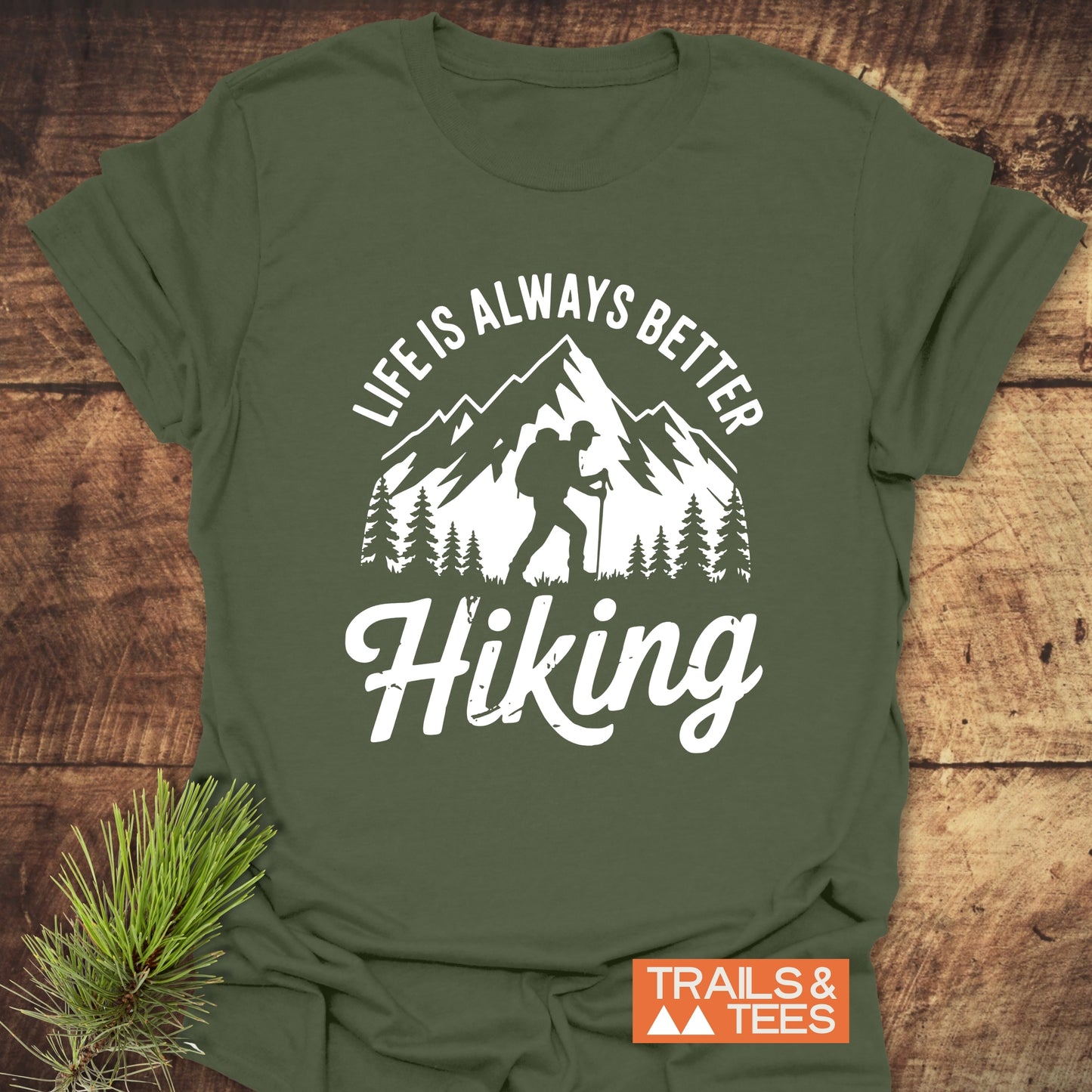 Forest-green Life Is Always Better Hiking T-shirt on wood surface, featuring a white backpacker graphic with mountains. Includes TRAILS & TEES logo.