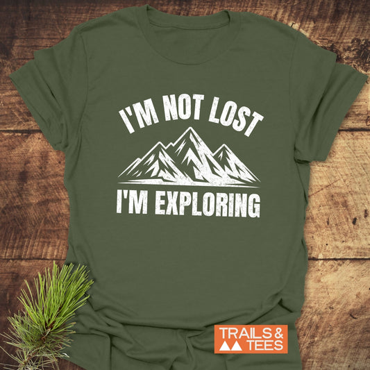 I'm Not Lost I'm Exploring T-Shirt with mountain graphic and logo, laid on wood with pine sprig, highlighting outdoor vibe and adventure-ready design.