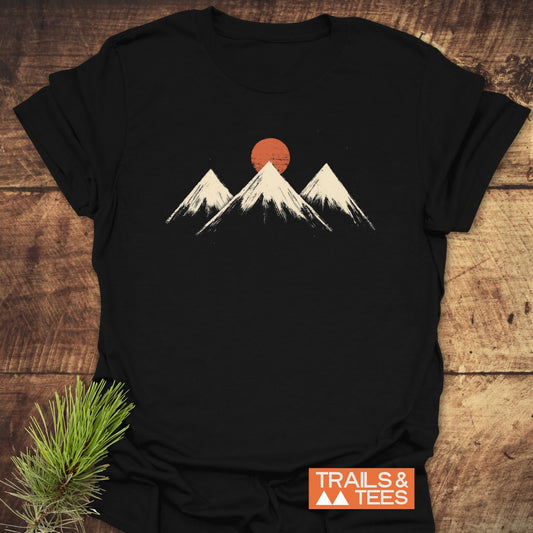 Three Mountains And Sun T-Shirt on wood, featuring mountain peaks with an orange sun. Beside it, a pine sprig and TRAILS & TEES logo.
