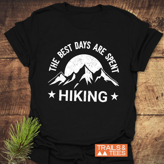 The Best Days Are Spent Hiking T-Shirt features mountain graphics and slogan, emphasizing outdoor adventure. Includes Trails & Tees logo and a pine sprig accent.
