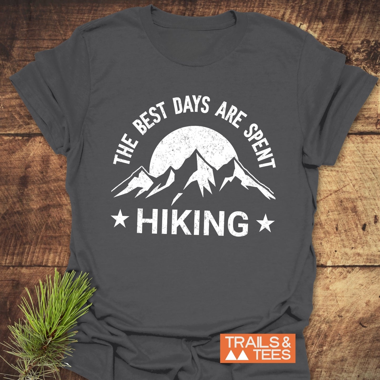 Charcoal-gray T-shirt with The Best Days Are Spent Hiking graphic, featuring a sun and mountains, laid on wood surface beside pine sprig.