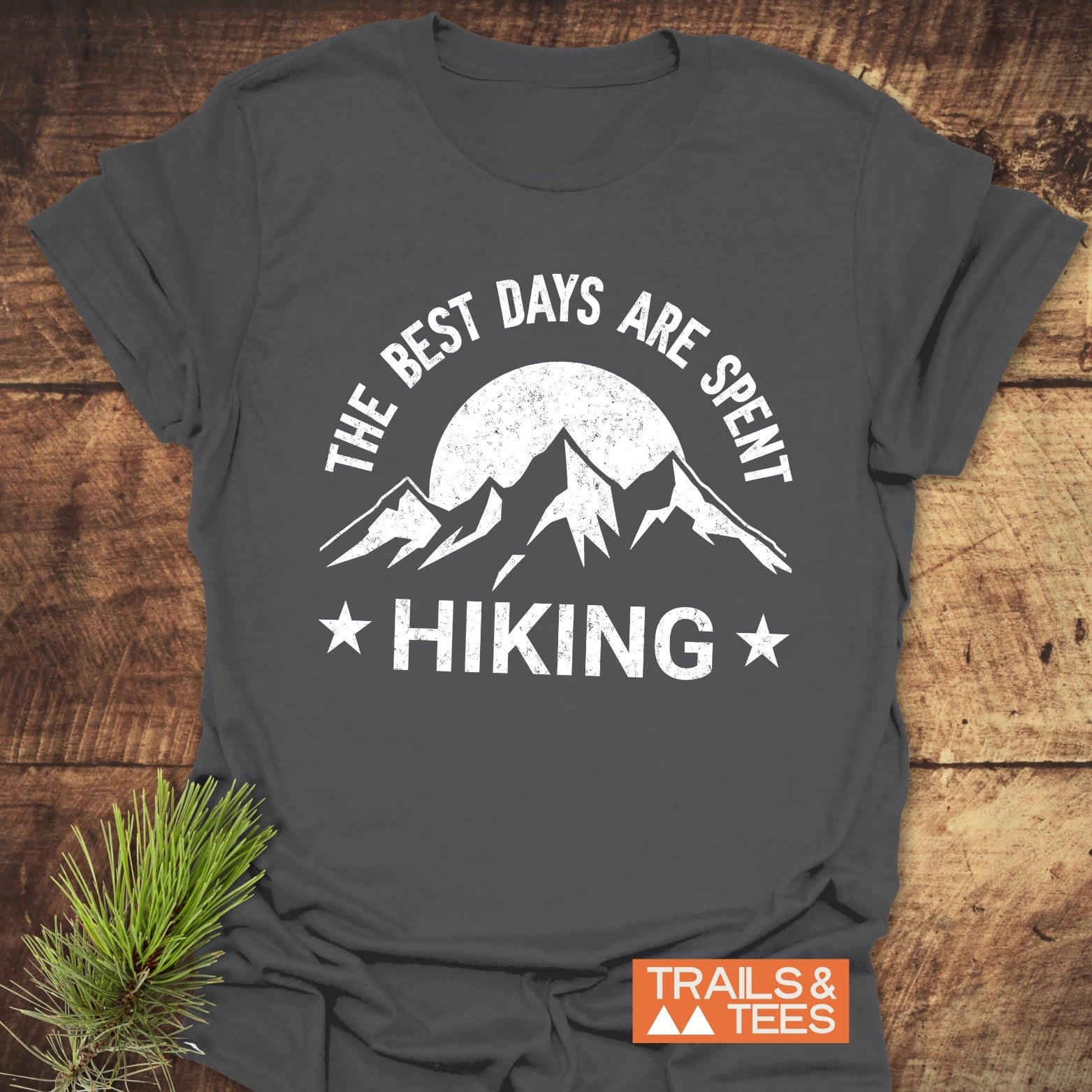 Charcoal-gray T-shirt with The Best Days Are Spent Hiking graphic, featuring a sun and mountains, laid on wood surface beside pine sprig.