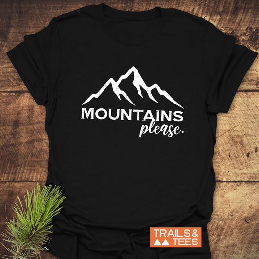 Black T-shirt with “MOUNTAINS please” and mountain line-drawing, branded “TRAILS & TEES,” laid on wood with pine sprig.