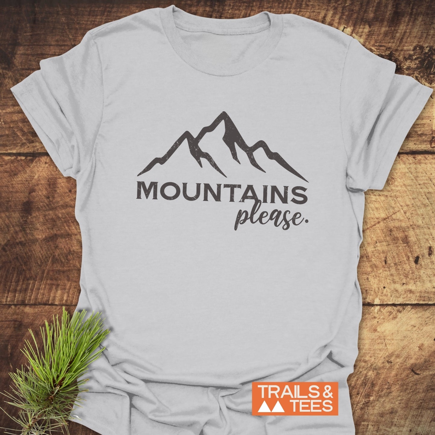 Mountains Please T-Shirt featuring mountain peak line-drawing and “MOUNTAINS please” text, styled with a pine sprig and Trails & Tees logo label.