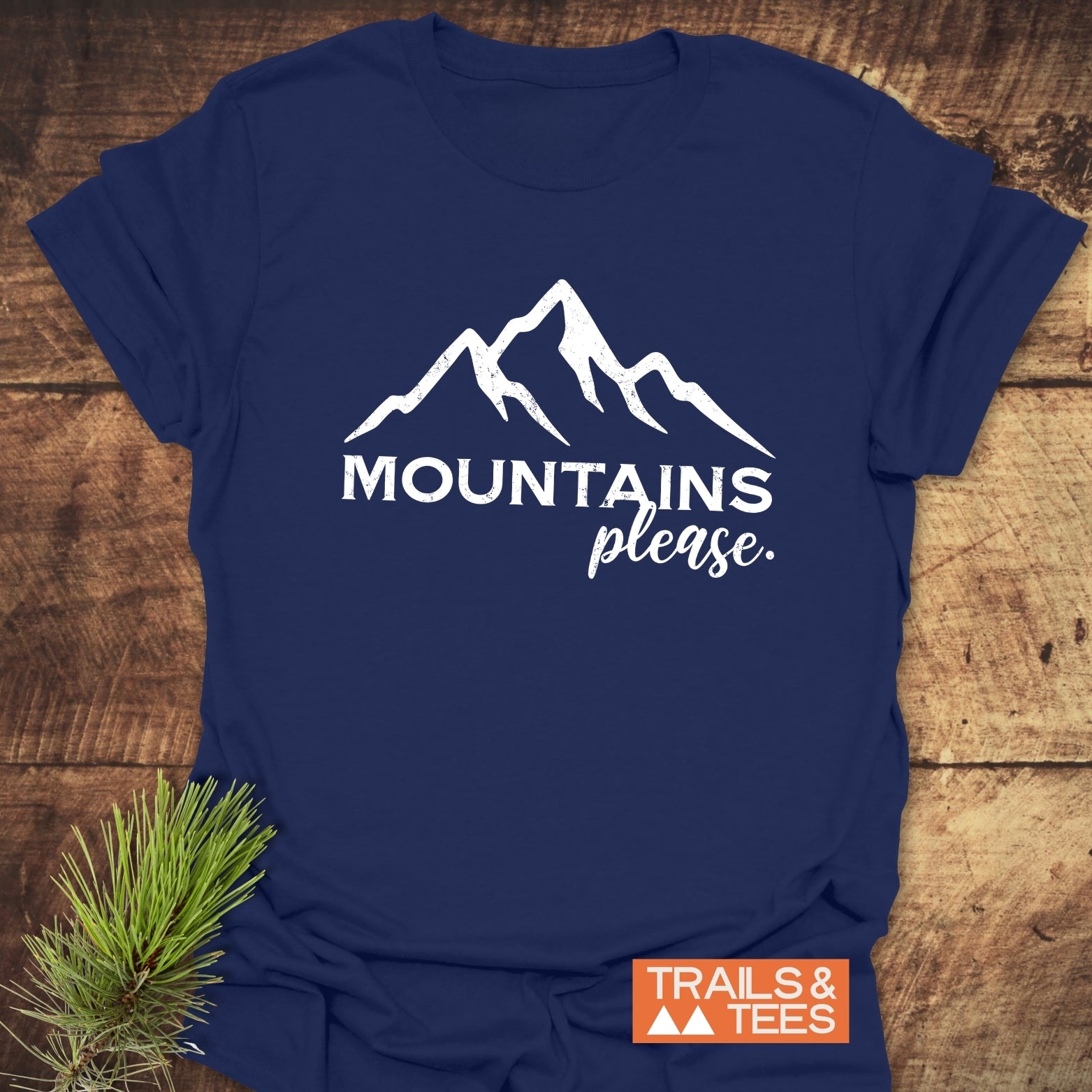 Navy-blue Mountains Please T-shirt with a white mountain silhouette, laid on wood, featuring an orange TRAILS & TEES logo and a pine branch accent.