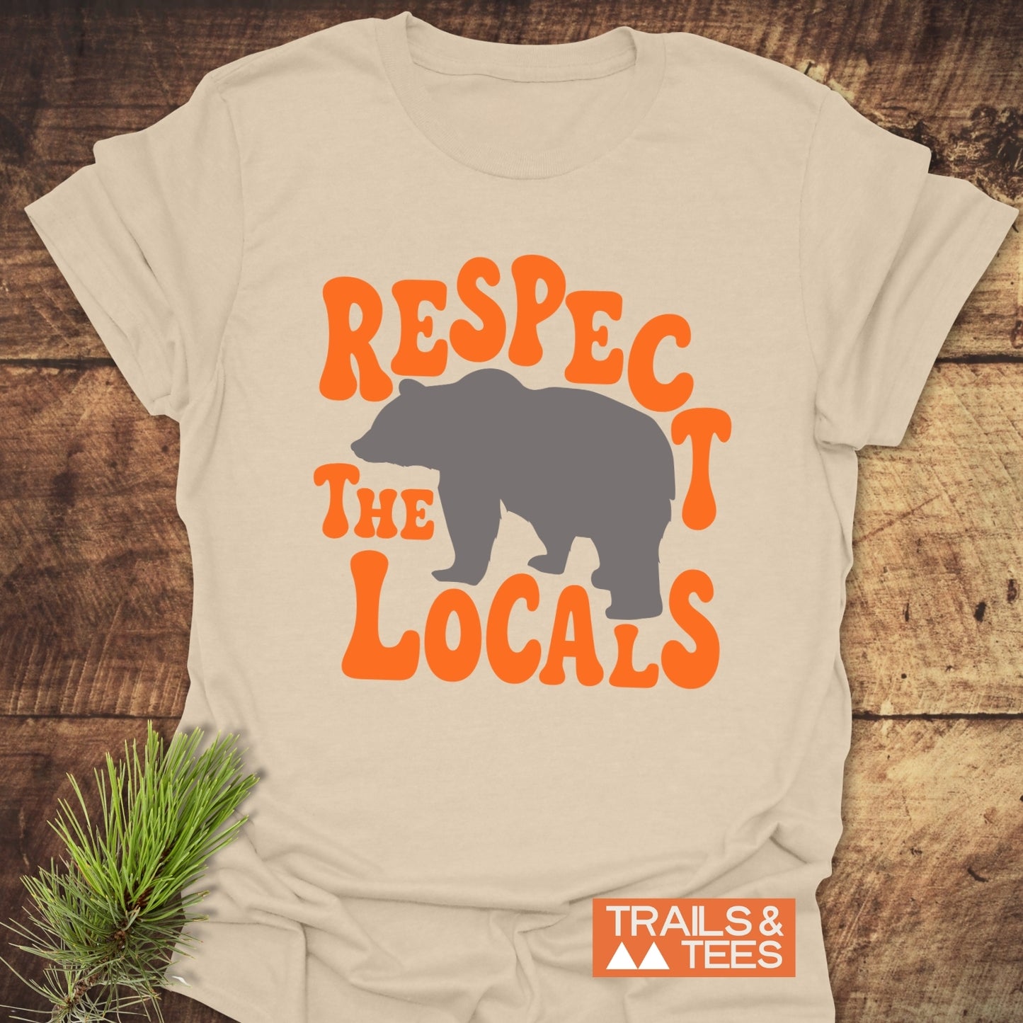 Respect The Locals Bear T-Shirt featuring a gray bear silhouette with bold orange lettering, laid flat with a pine sprig beside it.