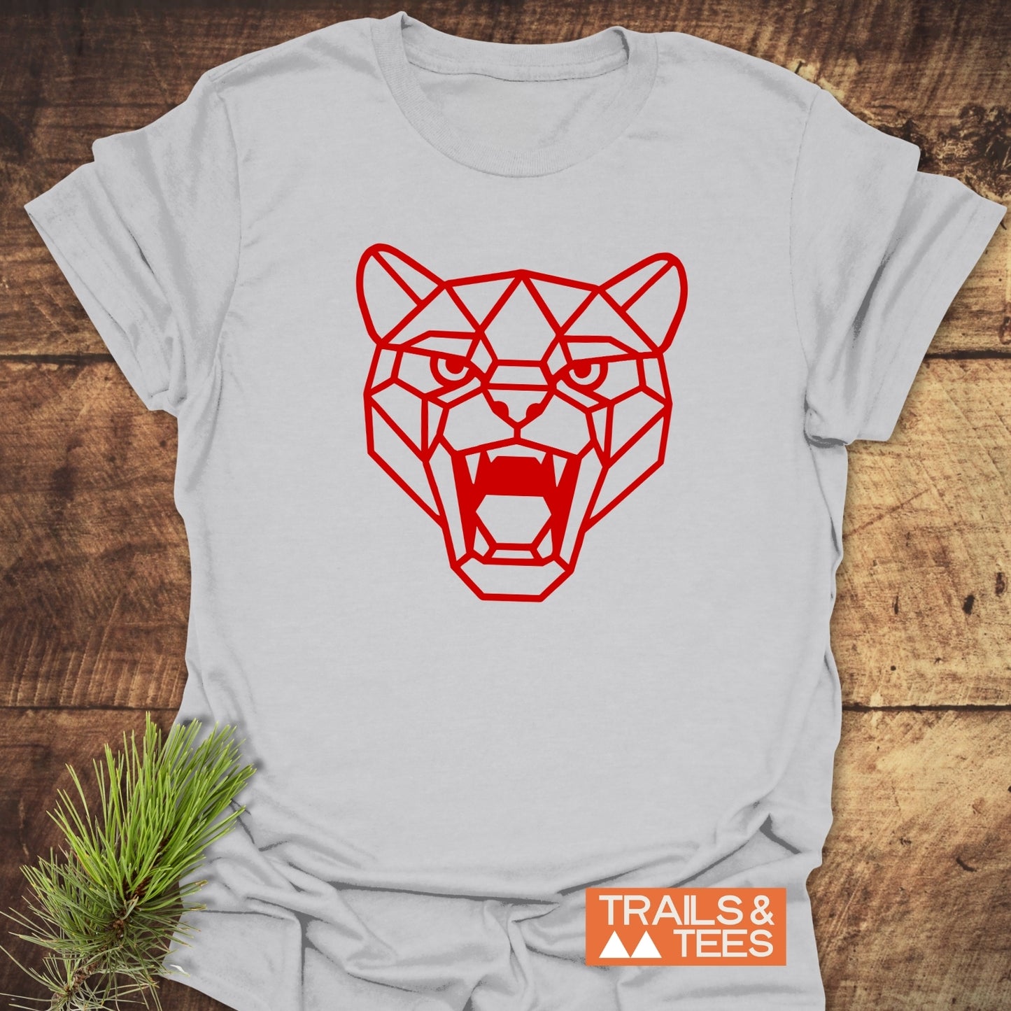 Geometric Cougar T-Shirt with a red big-cat design on light fabric, featuring a “TRAILS & TEES” label, laid on a wooden surface with pine sprig.