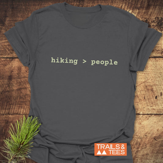 Hiking > People T-Shirt on wooden surface, featuring typewriter-style text, small TRAILS & TEES logo, and pine sprig, highlighting its outdoor-inspired design.