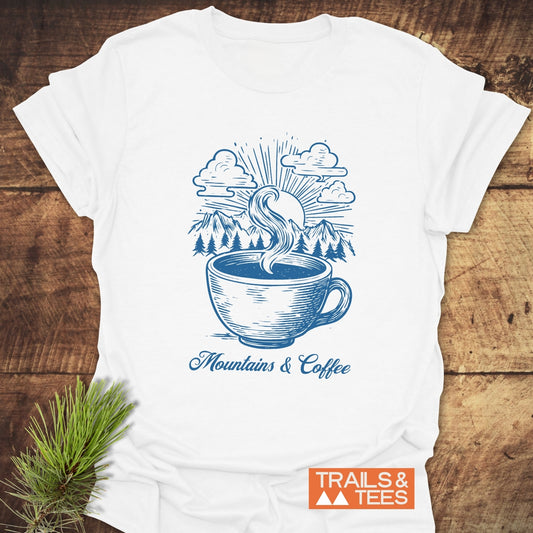 Mountains And Coffee T-Shirt featuring a blue coffee cup and mountain scene design, styled with an evergreen sprig, embodies outdoor adventure and comfort.