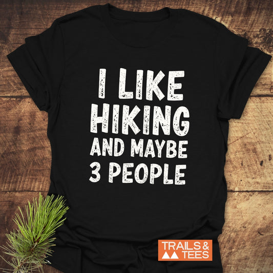 Black T-shirt with I LIKE HIKING AND MAYBE 3 PEOPLE in white letters, featuring a Trails & Tees logo, laid on wood with a pine sprig.