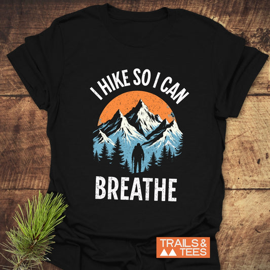 I Hike So I Can Breathe T-Shirt featuring mountain graphics, hiker silhouette, and slogan, laid on wood. Made from soft, durable cotton for outdoor enthusiasts.