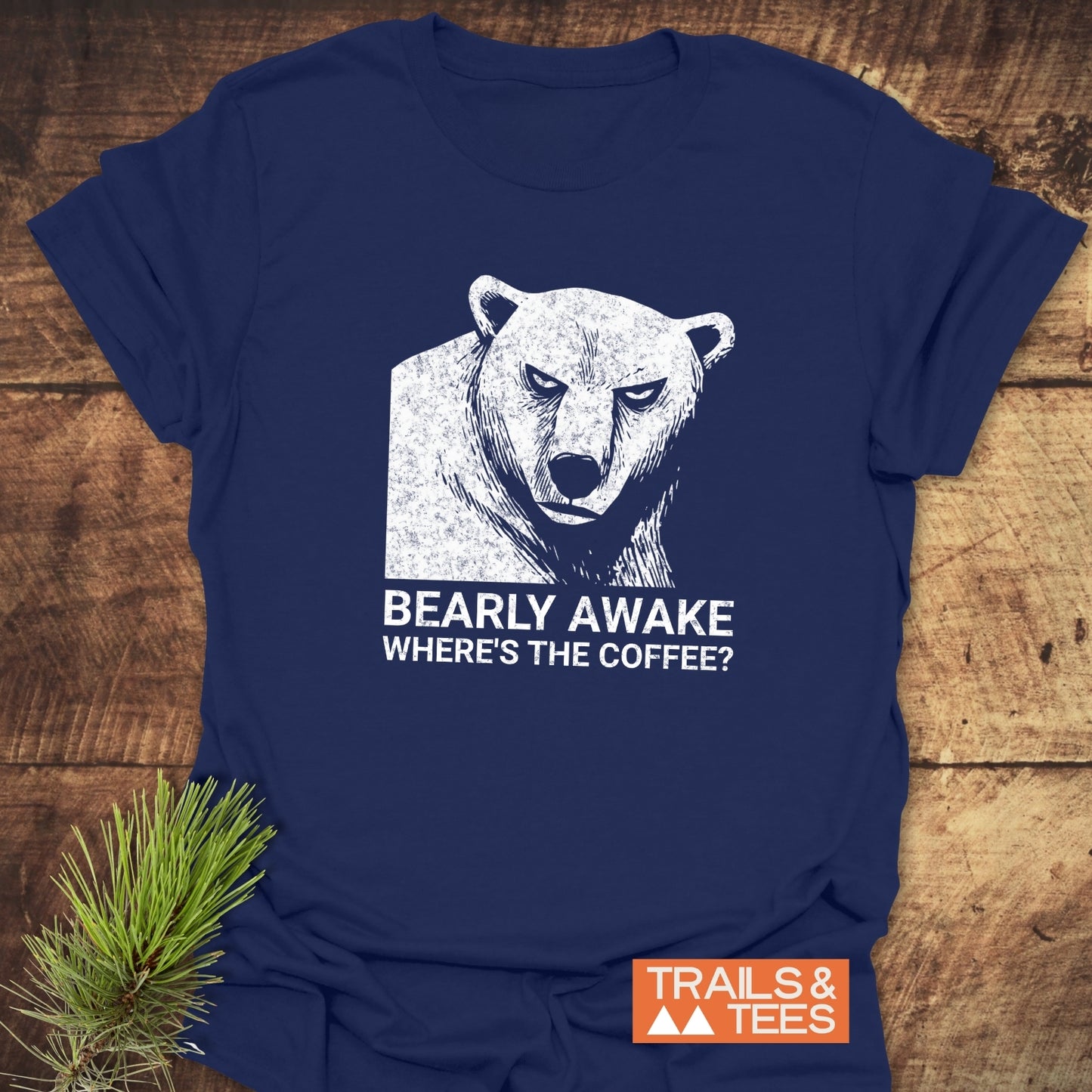 Bearly Awake T-Shirt with a grumpy bear sketch and slogan, crafted from soft cotton, laid on wood, featuring durable prints and Trails & Tees logo.