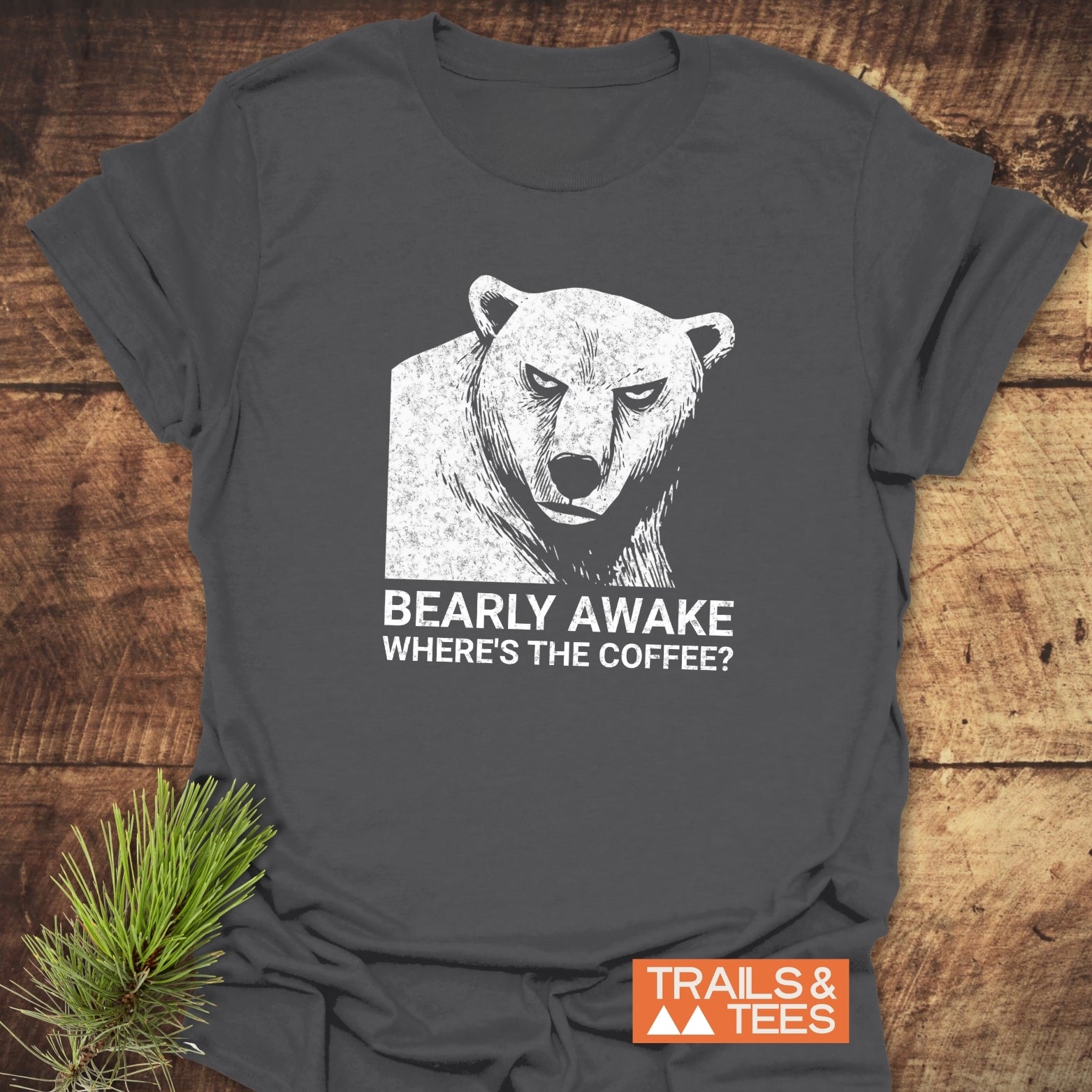 Bearly Awake T-Shirt featuring a distressed bear graphic and slogan on dark-gray cotton, laid on wood with a pine sprig beside it.