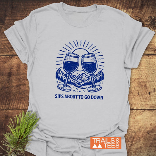 Sips About To Go Down T-Shirt featuring blue wine glass graphic on light fabric, laid on wood with pine bough; Trails & Tees logo visible.