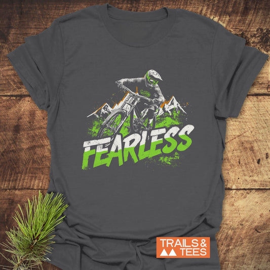 Fearless Mountainbiker T-Shirt featuring a downhill rider and mountain peaks, with FEARLESS text. Laid on wood surface, highlighting outdoor adventure spirit.