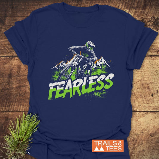 Fearless Mountainbiker T-Shirt on wooden surface, featuring a mountain biker design with mountains and FEARLESS text. Includes Trails & Tees logo.