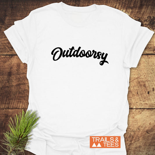 Outdoorsy T-Shirt featuring black script font on a white tee, with a pine sprig and TRAILS & TEES logo on a wood-plank background.