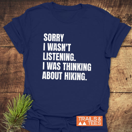 Navy-blue Sorry I Wasn't Listening Hiking T-Shirt with white slogan, featuring durable print and orange TRAILS & TEES logo, on wooden surface with pine sprig.