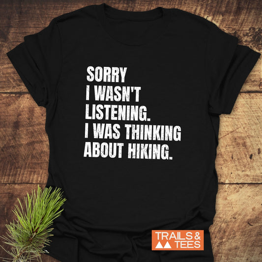 Sorry I Wasn't Listening Hiking T-Shirt with bold white text, laid on wood. Features soft ring-spun cotton and durable prints. 'TRAILS & TEES' logo visible.