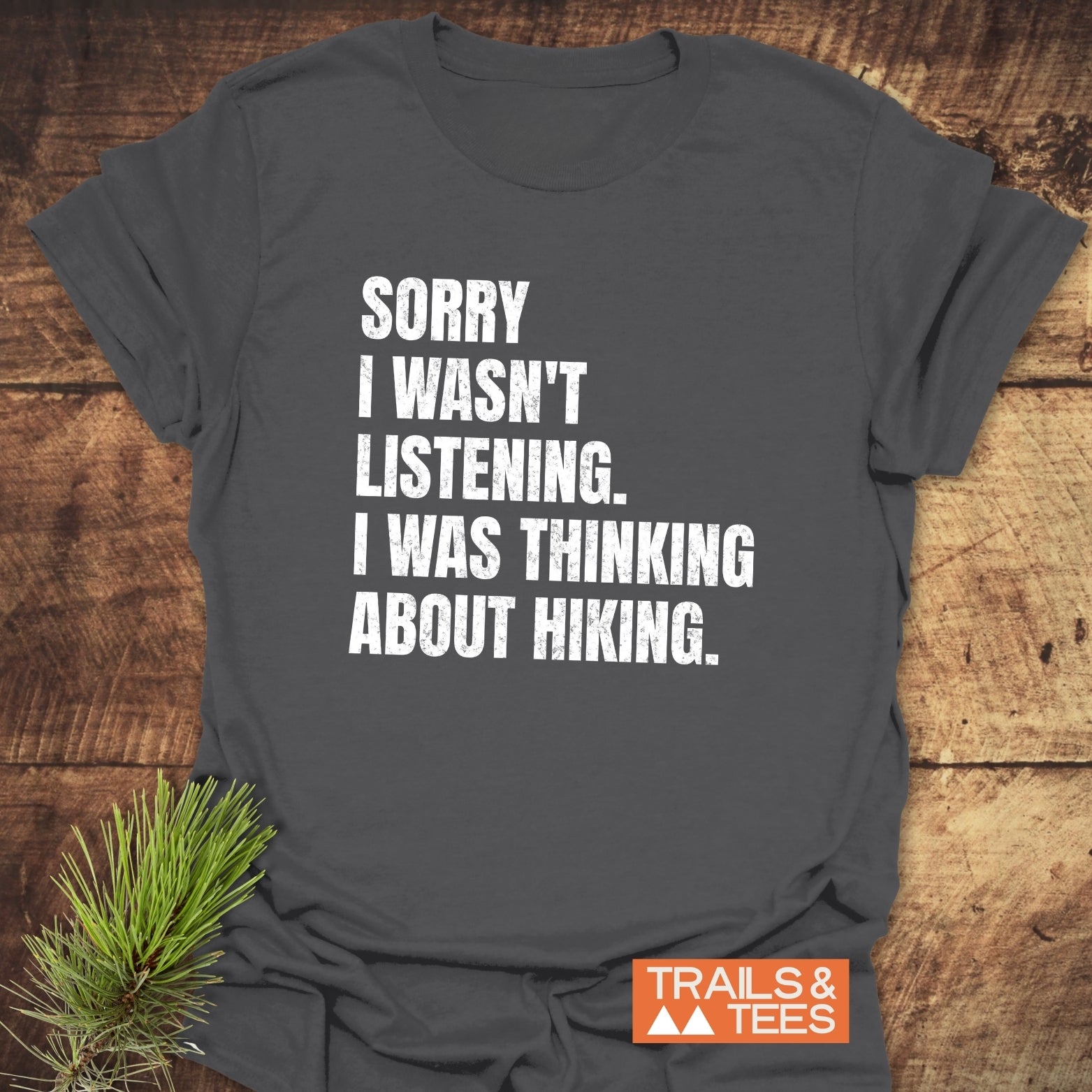 Charcoal-gray Sorry I Wasn't Listening Hiking T-Shirt on wood, featuring white text, orange TRAILS & TEES logo, highlighting comfort and adventure-ready design.