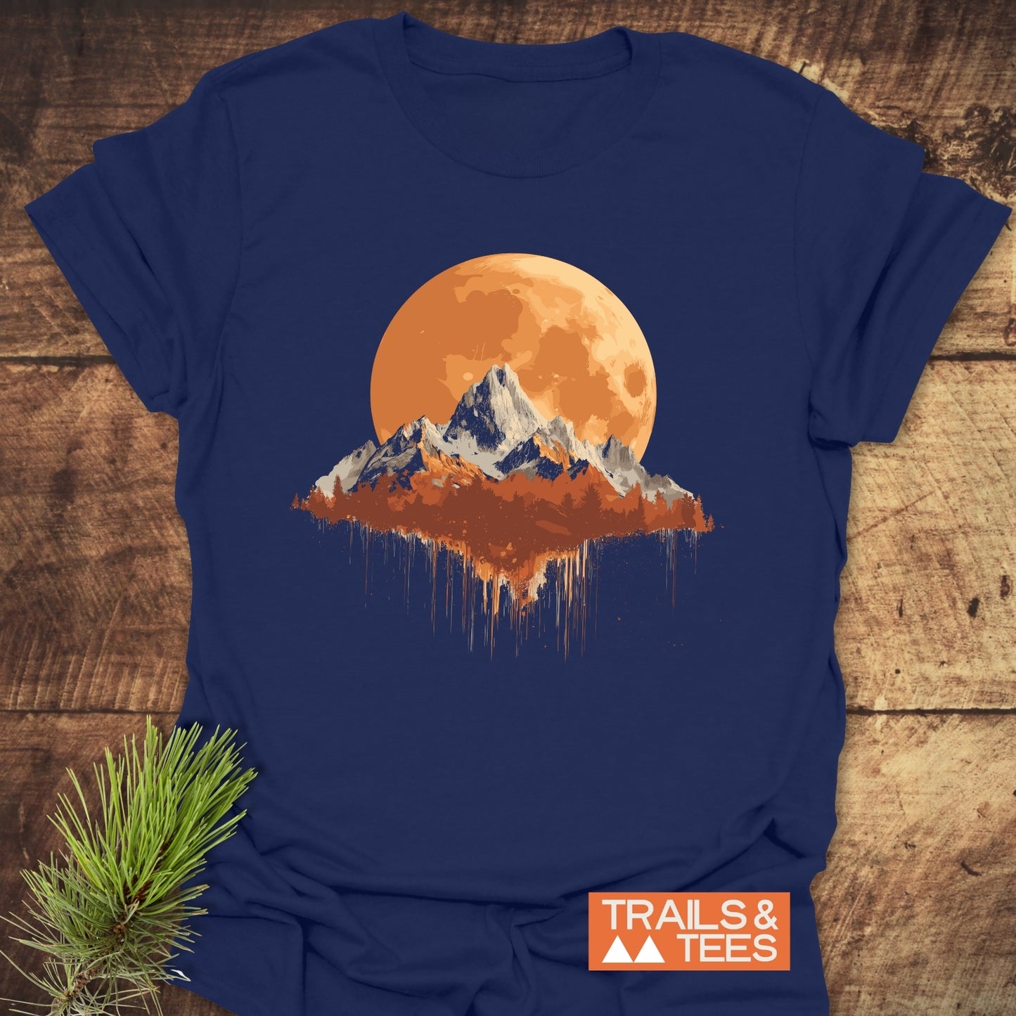 Orange Moon T-Shirt with mountain and moon graphic on navy fabric, featuring durable print and soft cotton, laid on rustic wood with pine sprig.