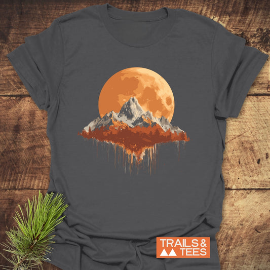 Orange Moon T-Shirt with stylized mountain scene printed on dark-gray fabric, showcasing a large orange moon. Includes TRAILS & TEES logo, laid on wood with pine sprig.