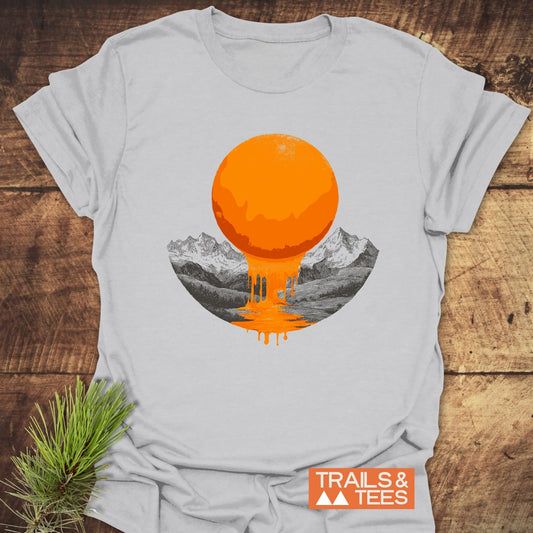 Melting Sun T-Shirt on rustic wood, featuring a dripping orange sun graphic over a lake and mountains, with TRAILS & TEES logo. Pine needles nearby.