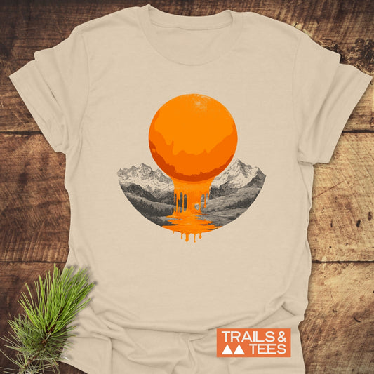 Melting Sun T-Shirt featuring a bright orange, dripping sun over sketched mountains; laid on a wooden surface with a pine sprig beside it.