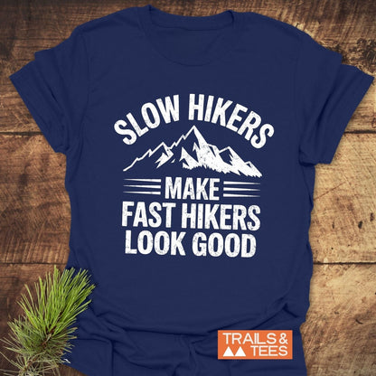 Navy-blue Slow Hikers T-Shirt with white mountain-themed text, laid on wood. Features orange label TRAILS & TEES and pine sprig beside it.