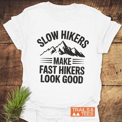 Slow Hikers T-Shirt featuring a mountain graphic and slogan, laid on wood with pine branch, emphasizes outdoor adventures. Includes “TRAILS & TEES” logo.
